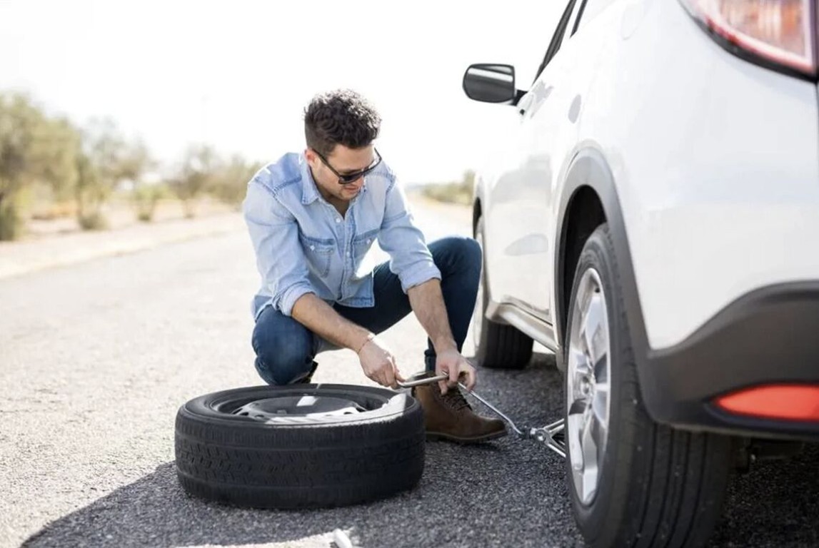 Man kneeling on the road changing a flat tire on a white SUV in Islip, NY